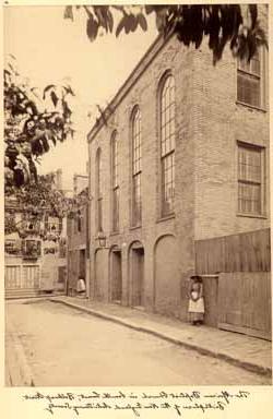 <p>Sepia photograph of a street and the side of a tall, brick building with four arched windows and doors and a large iron lamp. To the left of the building, a Black woman with a shawl, 帽子, 围裙, and long-sleeved dress stands against a wall with her hands clasped together in front of her.</p>