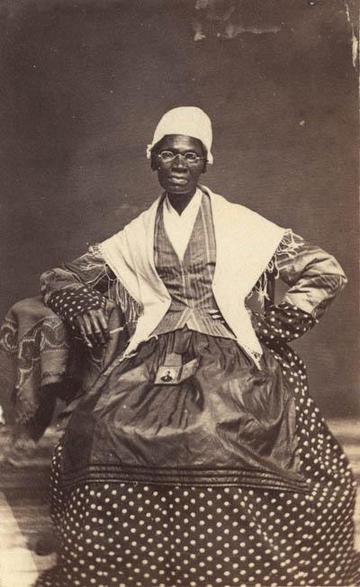 <p>Black and white photograph of a middle aged Black woman wearing glasses, a white cap and shawl, a polka-dotted dress, and a jacket and 围裙. She sits facing the camera with one hand on her hip and her other elbow resting on a small table. In her lap is a small framed photograph of her grandson.</p>